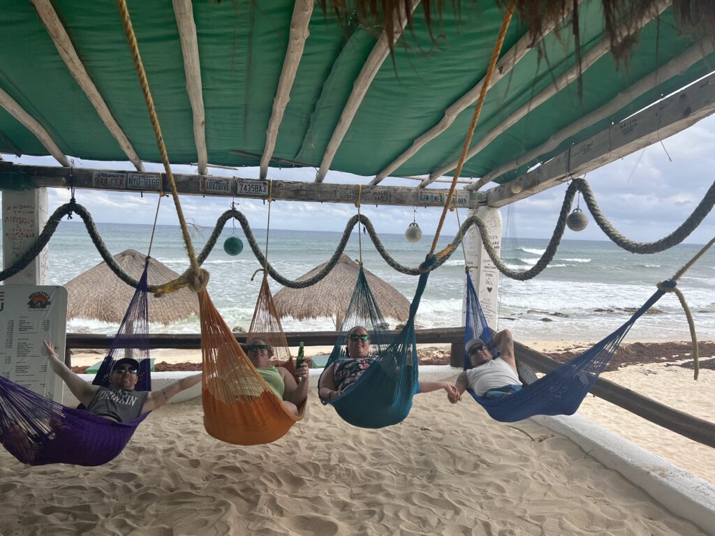 Four people relaxing in hammocks, with drinks in hand, in front of the ocean in Cozumel Mexico. The people are happy and posing for the camera. ultimate travel guide to cozumel