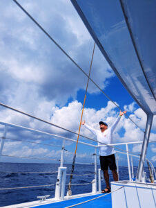 Adventure awaits! Short travel quotes for social media. Featured Image of a man on a catamaran boat, leaning against the rails at his waist with hands raised to the sky