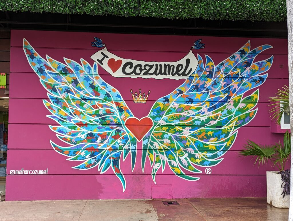 ultimate travel guide to cozumel. Popular Instagram Photo Spot, mural painting of angel wings, colorful on a magenta pink background, saying I love Cozumel across the top