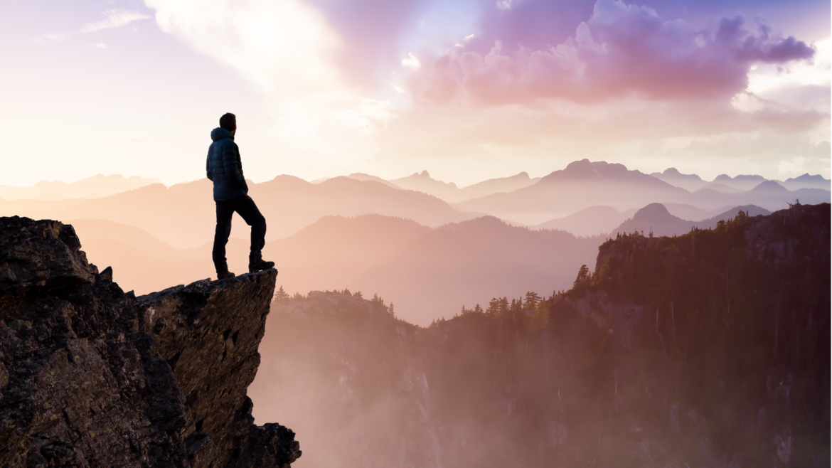 Silhouette of a climber standing on a rocky ledge overlooking misty mountain peaks at sunrise.